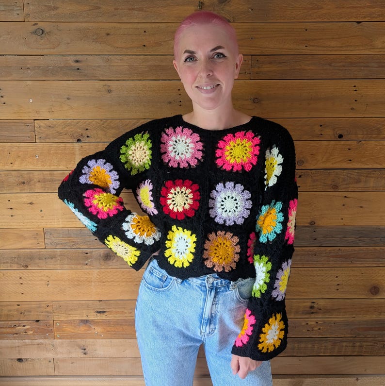 Puede incluir: Su&eacute;ter negro de crochet con cuadrados florales coloridos. El su&eacute;ter tiene cuello redondo y mangas largas con pu&ntilde;os acampanados. Las flores son en tonos rosa, amarillo, verde y naranja. La persona lleva vaqueros azul claro.