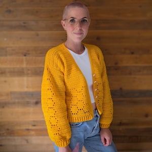 May include: A bright yellow, open-front crochet cardigan with a textured pattern and long sleeves. The cardigan is buttoned and worn over a white top and blue jeans. The background is a wooden wall.