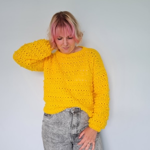 May include: A yellow crocheted sweater with a loose fit and a round neckline. The sweater has a textured stitch pattern and a slightly puffed sleeve.