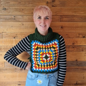 May include: A green turtleneck crochet vest with a colorful granny square design. The square features a pattern of yellow, blue, orange, and beige squares. The person is wearing a black and white striped long sleeve shirt and blue jeans.