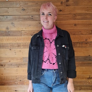 Puede incluir: Un suéter de cuello alto de crochet rosa con un diseño de encaje. El suéter se lleva sobre una chaqueta vaquera negra y unos vaqueros azules.