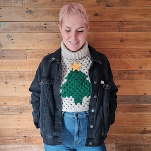 Può includere: Un maglione all'uncinetto fatto a mano con un motivo ad albero di Natale verde e una stella gialla, su uno sfondo grigio e bianco. Il maglione ha un collo alto e viene indossato con una giacca di jeans nera e jeans blu. Lo sfondo è una parete di legno.