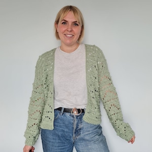 May include: A light green crocheted cardigan with a delicate open stitch pattern. The cardigan has a V-neck and long sleeves. The model is wearing the cardigan over a white t-shirt and blue jeans.