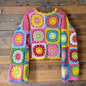 Puede incluir: Un colorido su&eacute;ter de crochet hecho a mano con un dise&ntilde;o de patchwork de cuadrados florales en tonos rosa, amarillo, azul, verde y rojo. El su&eacute;ter tiene mangas largas y un escote festoneado, perfecto para un look bohemio &uacute;nico.