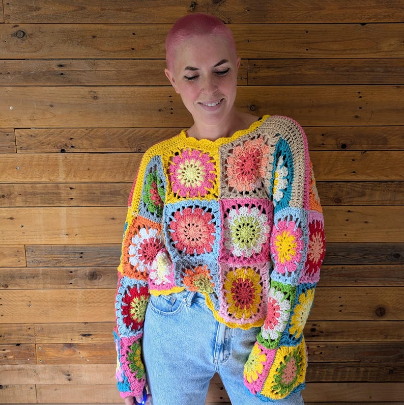 Puede incluir: Un colorido su&eacute;ter de ganchillo con un dise&ntilde;o de patchwork de cuadrados florales en tonos de amarillo, rosa, azul y verde. El su&eacute;ter tiene mangas largas y un escote festoneado. La prenda se usa con jeans azules.