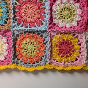 Puede incluir: Una colorida manta de ganchillo hecha a mano con un dise&ntilde;o de patchwork. Los cuadrados muestran patrones florales en tonos de rosa, naranja, amarillo y azul, con un borde amarillo festoneado. La manta est&aacute; hecha de hilo suave.