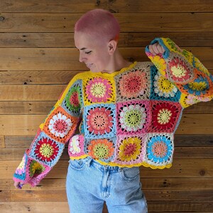 Puede incluir: Un su&eacute;ter de ganchillo hecho a mano y colorido, con paneles cuadrados en varios colores, incluyendo amarillo, rosa, azul y verde. El su&eacute;ter tiene mangas largas y un escote festoneado. El dise&ntilde;o muestra un patr&oacute;n floral dentro de cada cuadrado.