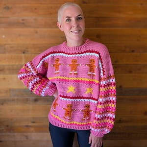May include: A pink crochet sweater with gingerbread man and star designs. The sweater features a round neck and long sleeves with a mix of pink, white, yellow, and orange colors. The gingerbread men are orange with white accents.