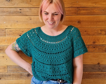 May include: A teal crochet top with a delicate openwork pattern. The top has short sleeves and a round neckline. The model is wearing the top with blue jeans.
