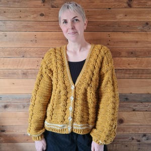 May include: A mustard yellow crocheted cardigan with a cable knit pattern and white trim. The cardigan has a V-neck and button closure.