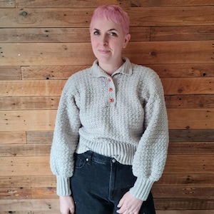 Puede incluir: Un suéter de crochet gris con cuello y cierre de botones. El suéter tiene un patrón de punto texturizado y mangas largas.