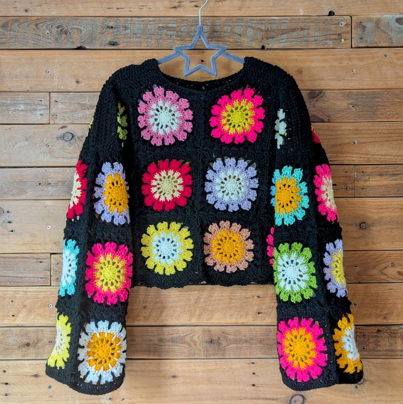 Puede incluir: Su&eacute;ter de crochet negro con cuadrados florales coloridos. El su&eacute;ter tiene cuello redondo y mangas largas. Las flores son en tonos rosa, amarillo, azul y blanco. El su&eacute;ter est&aacute; colgado de una percha en forma de estrella sobre un fondo de madera.