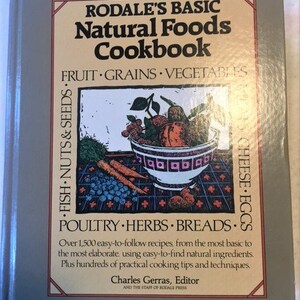 May include: A gray cookbook with the title "Rodale's Basic Natural Foods Cookbook" and a subtitle "Fruit Grains Vegetables". The cover features a bowl of fruit and vegetables with a colorful illustration of various foods.