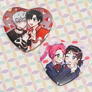 May include: Two heart-shaped buttons featuring anime-style illustrations. One button shows two male figures, one with silver hair and the other in a red jacket. The other button depicts two female figures with pink hair and glasses.