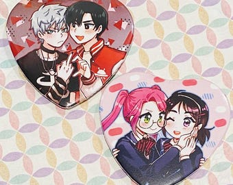 May include: Two heart-shaped buttons featuring anime-style illustrations. One button shows two male figures, one with silver hair and the other in a red jacket. The other button depicts two female figures with pink hair and glasses.