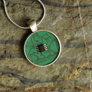 Puede incluir: Un collar de cadena de plata con un colgante redondo que presenta una placa de circuito verde con un chip de circuito integrado negro en el centro.