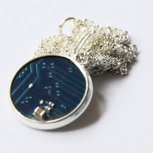 Puede incluir: Un collar de cadena de plata con un colgante redondo que presenta un diseño de placa de circuito azul.