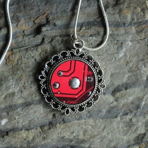 Puede incluir: Un collar de cadena de plata con un colgante redondo que presenta un diseño de placa de circuito rojo encerrado en un marco de filigrana de plata.