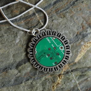 Puede incluir: Un collar de cadena de plata con un colgante redondo que presenta una placa de circuito verde dentro de un marco de filigrana de plata. El colgante tiene el texto "POWER" y "144" impreso en él.