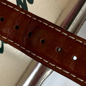 K&ouml;nnte beinhalten: Nahaufnahme eines braunen Lederg&uuml;rtels mit wei&szlig;en N&auml;hten und mehreren gleichm&auml;&szlig;ig verteilten L&ouml;chern. Der G&uuml;rtel scheint abgenutzt zu sein, mit einem leicht abgenutzten Aussehen. Die N&auml;hte sind sauber und kontrastieren mit dem dunkleren Leder.