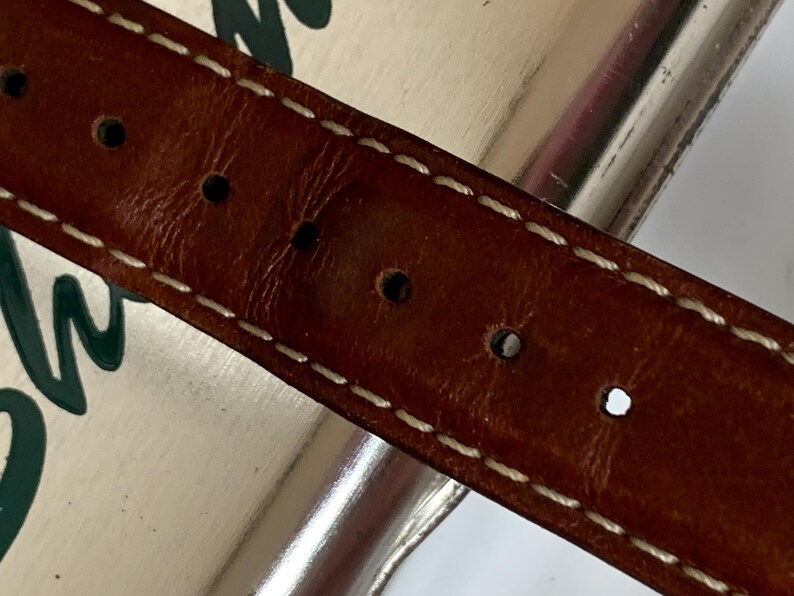 K&ouml;nnte beinhalten: Nahaufnahme eines braunen Lederg&uuml;rtels mit wei&szlig;en N&auml;hten und mehreren gleichm&auml;&szlig;ig verteilten L&ouml;chern. Der G&uuml;rtel scheint abgenutzt zu sein, mit einem leicht abgenutzten Aussehen. Die N&auml;hte sind sauber und kontrastieren mit dem dunkleren Leder.