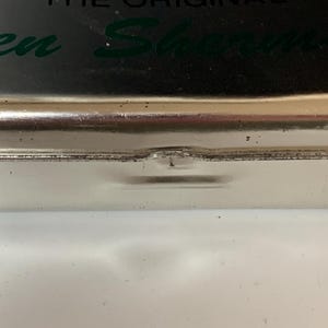 K&ouml;nnte beinhalten: Nahaufnahme eines silbernen Metallbeh&auml;lters mit dem gr&uuml;nen Schriftzug "Ben Sherman". Der Beh&auml;lter hat eine leicht eingedellte Kante und liegt auf einer wei&szlig;en Oberfl&auml;che.