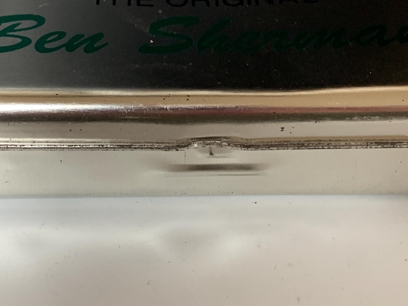 K&ouml;nnte beinhalten: Nahaufnahme eines silbernen Metallbeh&auml;lters mit dem gr&uuml;nen Schriftzug "Ben Sherman". Der Beh&auml;lter hat eine leicht eingedellte Kante und liegt auf einer wei&szlig;en Oberfl&auml;che.