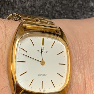 Puede incluir: Un reloj Timex Quartz dorado con esfera blanca y manecillas e indicadores negros. El reloj tiene una esfera rectangular con esquinas redondeadas y una correa de metal dorado. La palabra "TIMEX" está impresa en la parte superior y "QUARTZ" en la inferior.