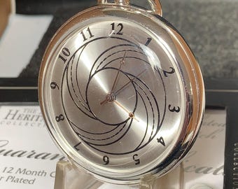 May include: Silver-toned pocket watch with a unique design on the face. The watch features a silver dial with black markings and a distinctive spiral pattern. The watch is displayed on a clear stand, showcasing its intricate details and craftsmanship.