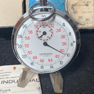 Może przedstawiać: Zegarek stopera Leonidas w stylu vintage z białą tarczą, czarnymi wskazówkami i czerwonymi cyframi. Stoper ma srebrną metalową obudowę i pierścień na górze. Tekst "LEONIDAS 7 JEWELS" jest wydrukowany na tarczy. Stoper jest wyświetlany na przezroczystym stojaku.