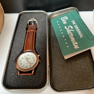K&ouml;nnte beinhalten: Eine Ben Sherman Uhr mit braunem Lederarmband und kupferfarbenem Geh&auml;use, pr&auml;sentiert in einer Metalldose. Das Zifferblatt ist wei&szlig; mit schwarzen Ziffern. Die Dose enth&auml;lt eine Garantiekarte.