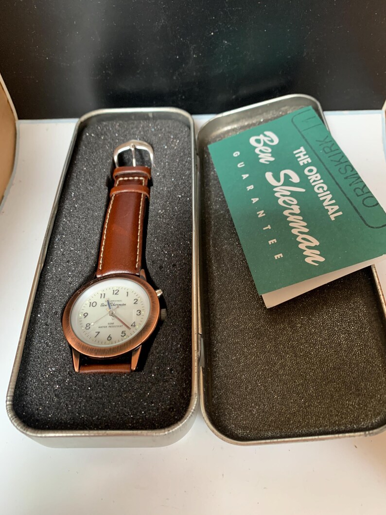 K&ouml;nnte beinhalten: Eine Ben Sherman Uhr mit braunem Lederarmband und kupferfarbenem Geh&auml;use, pr&auml;sentiert in einer Metalldose. Das Zifferblatt ist wei&szlig; mit schwarzen Ziffern. Die Dose enth&auml;lt eine Garantiekarte.