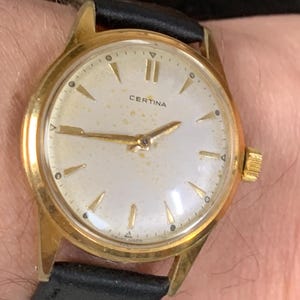 Op de afbeelding: Een gouden polshorloge met een witte wijzerplaat en een zwarte leren band. De wijzerplaat heeft zwarte Romeinse cijfers als uurmarkeringen en een zwarte secondewijzer. Op de wijzerplaat staat ook het woord "Certina" gedrukt.
