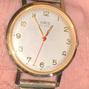 Puede incluir: Un reloj de pulsera dorado con una esfera blanca y marcadores de hora con números romanos negros. El reloj tiene una correa dorada y dice "ORIS ANTI-SHOC" en la esfera. "SWISS MADE" está impreso en la esfera.
