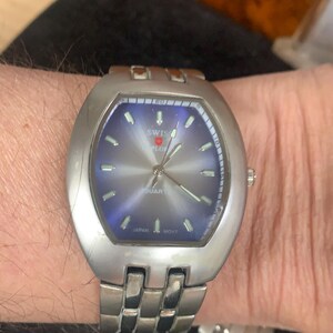 May include: A silver wristwatch with a blue face and a silver band. The watch face has the words "Swiss Pilot Quartz" and "Japan Movt" printed on it.