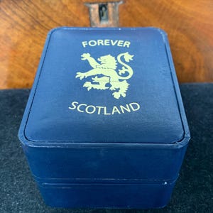 Puede incluir: Una caja azul marino con letras doradas y un emblema dorado. El texto en la caja dice "FOREVER SCOTLAND". La caja es rectangular y parece estar hecha de un material similar al cuero.