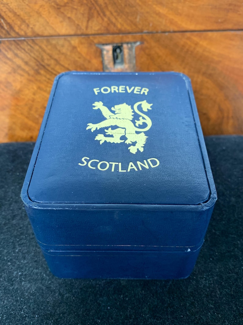 Puede incluir: Una caja azul marino con letras doradas y un emblema dorado. El texto en la caja dice "FOREVER SCOTLAND". La caja es rectangular y parece estar hecha de un material similar al cuero.