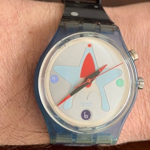 Puede incluir: Un reloj Swatch azul y blanco con un diseño de estrella roja y blanca en la esfera. El reloj tiene una correa negra con lunares blancos.