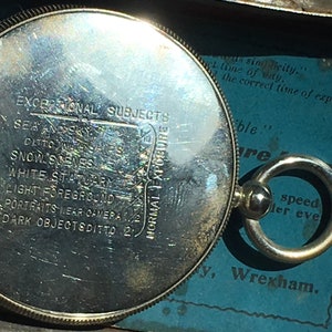 Op de afbeelding: Een zilverkleurige, ronde fotografische lichtmeter met gegraveerde tekst, waaronder "EXCEPTIONAL SUBJECTS" en "NORMAL EXPOSURE". Het heeft een ring om op te hangen. De achtergrond is blauw met extra tekst. Een vintage item.