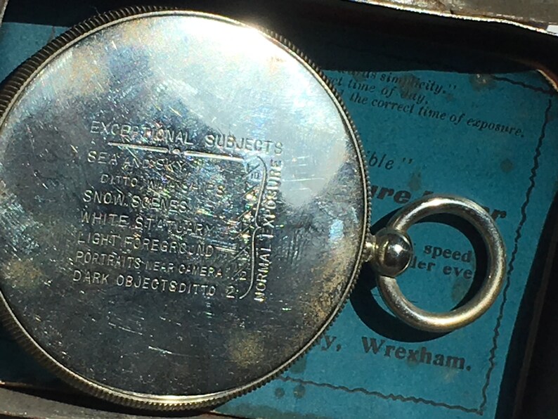 Op de afbeelding: Een zilverkleurige, ronde fotografische lichtmeter met gegraveerde tekst, waaronder "EXCEPTIONAL SUBJECTS" en "NORMAL EXPOSURE". Het heeft een ring om op te hangen. De achtergrond is blauw met extra tekst. Een vintage item.