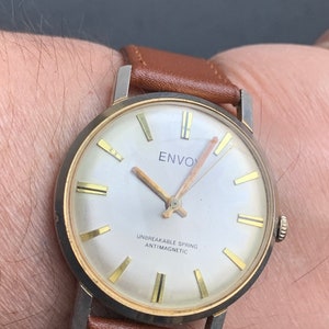 Puede incluir: Un reloj de pulsera vintage con caja plateada, detalles dorados y correa de cuero marrón. La esfera blanca muestra la palabra "ENVOY" y el texto "UNBREAKABLE SPRING ANTIMAGNETIC".
