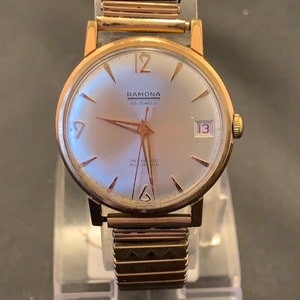 May include: A vintage Ramona wristwatch with a gold-tone case and a silver dial. The watch features gold hands and hour markers, a date display at the 3 o'clock position, and a gold-tone expansion band. The watch face reads "RAMONA 25 JEWELS" and "INCA-ROC AUTOMATIC".