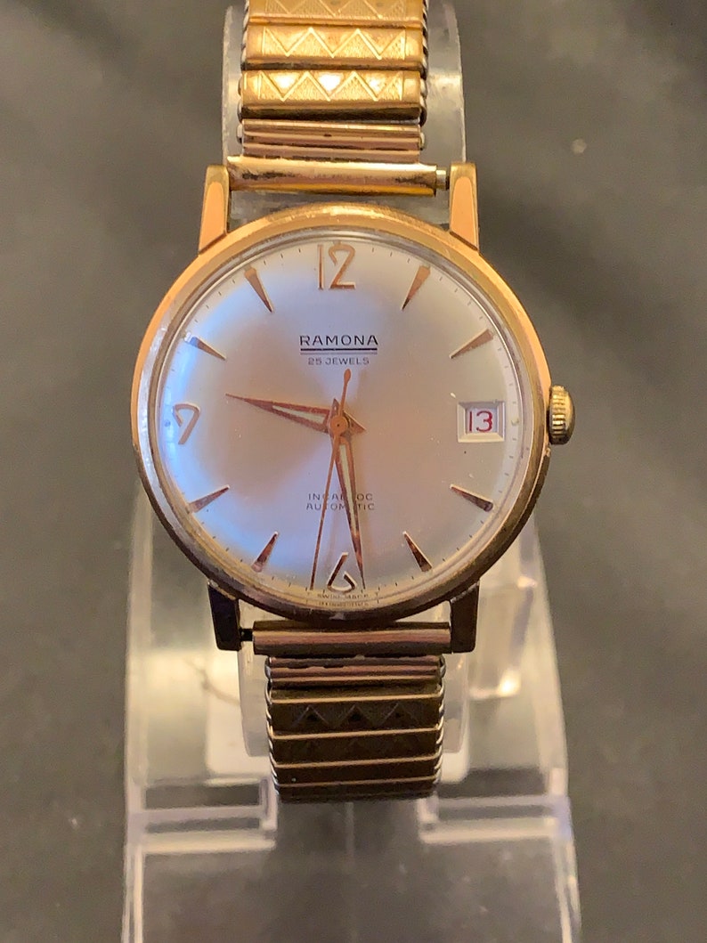 May include: A vintage Ramona wristwatch with a gold-tone case and a silver dial. The watch features gold hands and hour markers, a date display at the 3 o'clock position, and a gold-tone expansion band. The watch face reads "RAMONA 25 JEWELS" and "INCA-ROC AUTOMATIC".