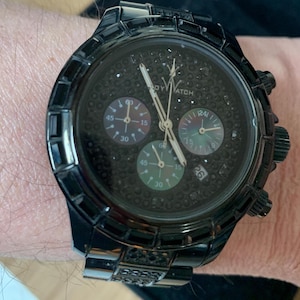 May include: A black wristwatch with a textured face and multiple sub-dials. The watch has a black bezel and strap, with the brand name "ROY WATCH" visible. The watch is worn on a wrist.