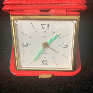 May include: A red leather-covered alarm clock with a gold-tone frame and a white face. The clock has a green second hand and the text "ELO 7 Jewels" on the face.