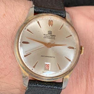 May include: A gold-tone wristwatch with a white face and black leather band. The watch face has black hands, black Roman numeral hour markers, and a date window at the 3 o'clock position. The watch face also has the text "Multy Prima Superflat 21 Datomatic" and "Antimagnetic".