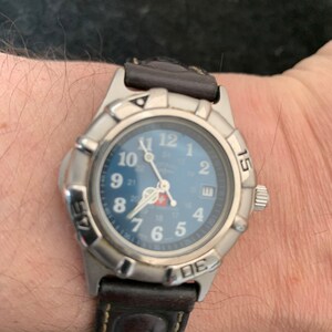 May include: A silver wristwatch with a blue face and a brown leather strap. The watch features white numerals and hands, and a date display. The bezel has markings at the 15 and 45-minute positions. The strap is brown leather.