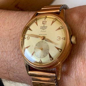 May include: A vintage gold-toned wristwatch with a cream-colored face. The watch features the brand name "Codhor" and a small seconds dial. The watch has gold-toned hands and hour markers, and a gold-toned metal band.