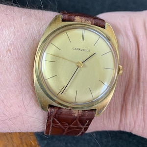 May include: A gold-toned wristwatch with a brown leather band. The watch face is round and has a gold-colored dial with black hands and Roman numeral hour markers. The word "Caravelle" is printed in black on the dial.