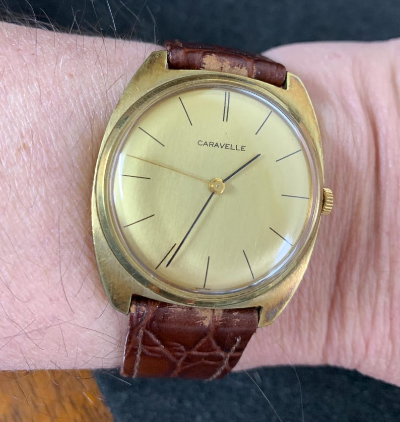 May include: A gold-toned wristwatch with a brown leather band. The watch face is round and has a gold-colored dial with black hands and Roman numeral hour markers. The word "Caravelle" is printed in black on the dial.